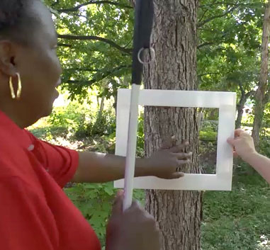 a woman is reaching through a picture frame which is held up to a tree trunk. She is tracing the side of the tree trunk to see which part would be on the picture plane and what part of the trunk would be off the picture plane.